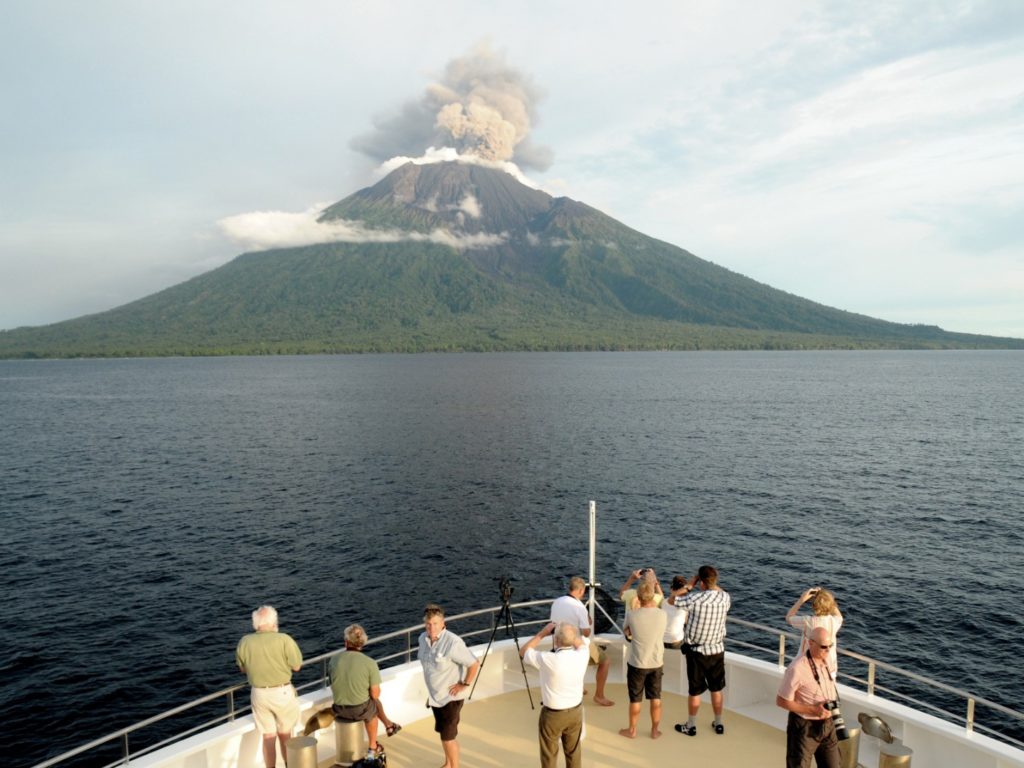 Bow of cruise boat with passengers looking towards snoking conical volcano island,