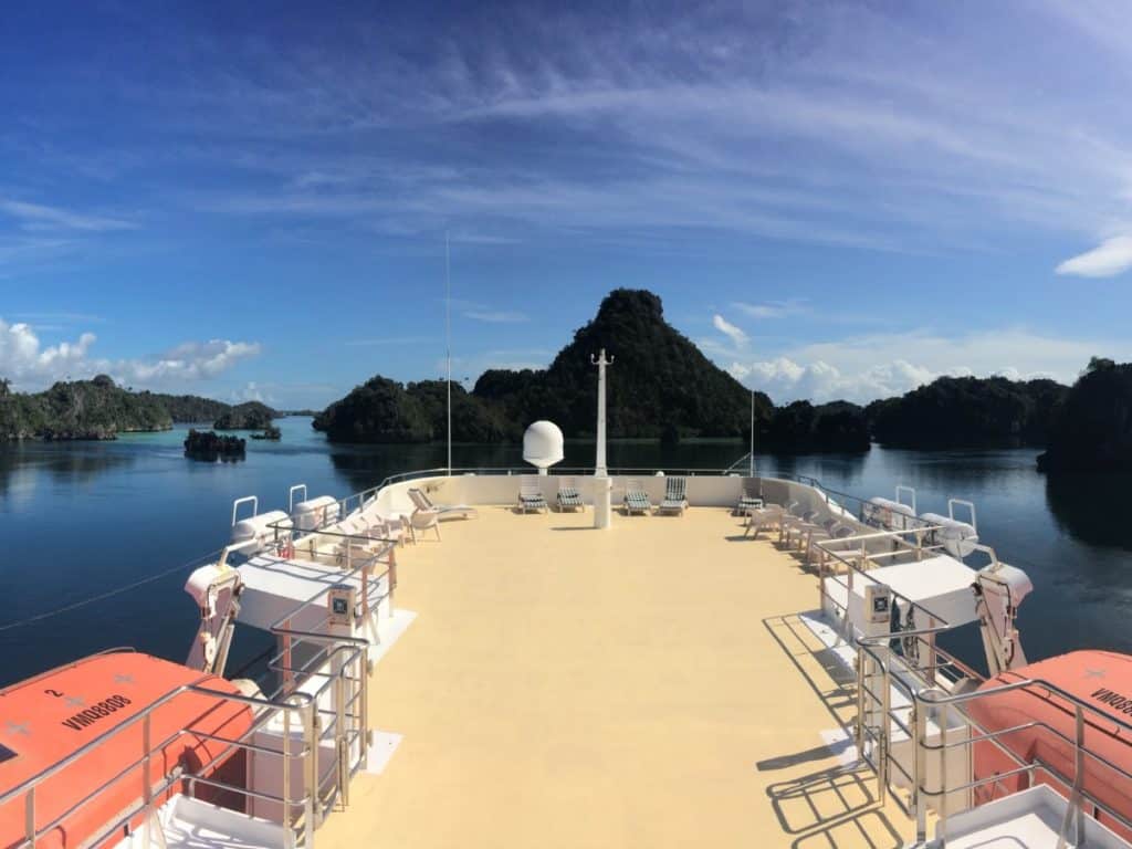 Stern of boat cruising with views of sillhouetted island peaks on calm blue water.