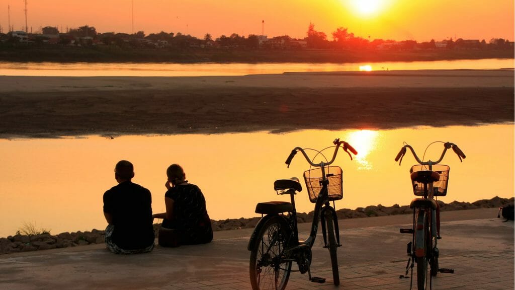Silhouetted couple sat on the banks of the Mekong at sunset with their bikes.