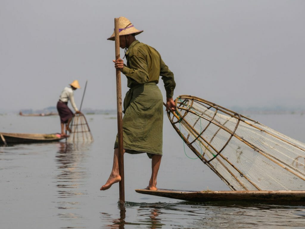 Fishermen on water stood on traditional wooden fishing canoe on one leg with conical shaped fishing net.