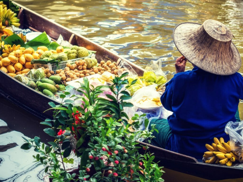 Side shot of floating market trader wearing traditional wicker hat with boat full of colourful fruit and veg.