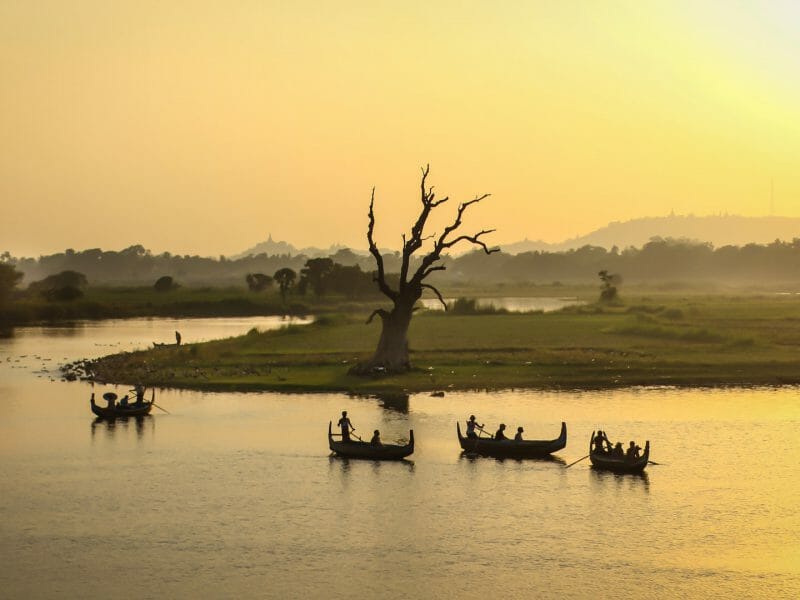 River at sunset with silhouettes of four traditional boats against yellow sky.