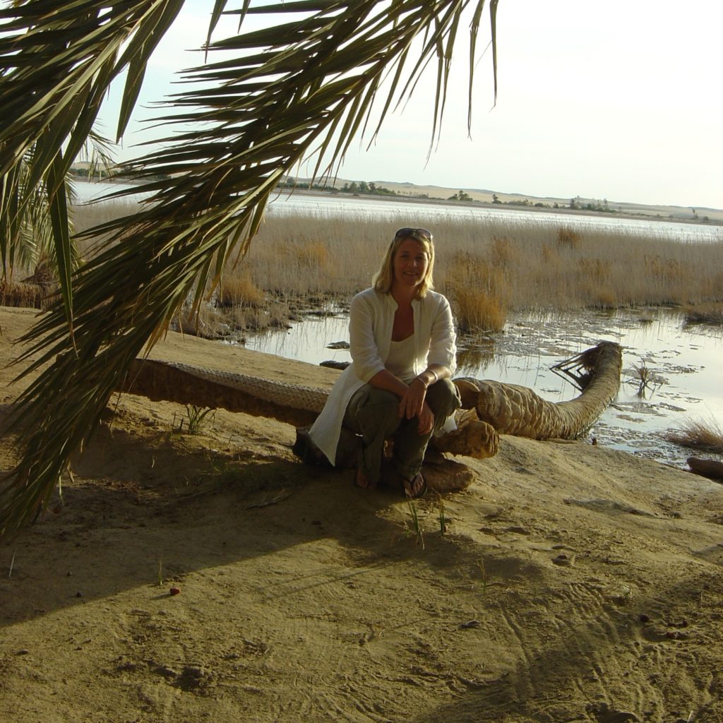 Kate sat on log under palm tree against backdrop of lake and western desert.