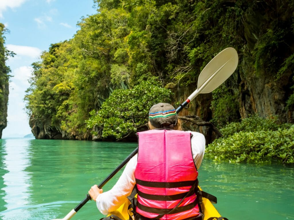 Rear of person in red lifejacket kayaking on emerald water surrounded by jungle covered cliffs.