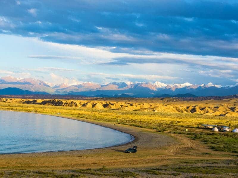 View of lime green grassland leading to shores of curved lake and snowy distant mountains.