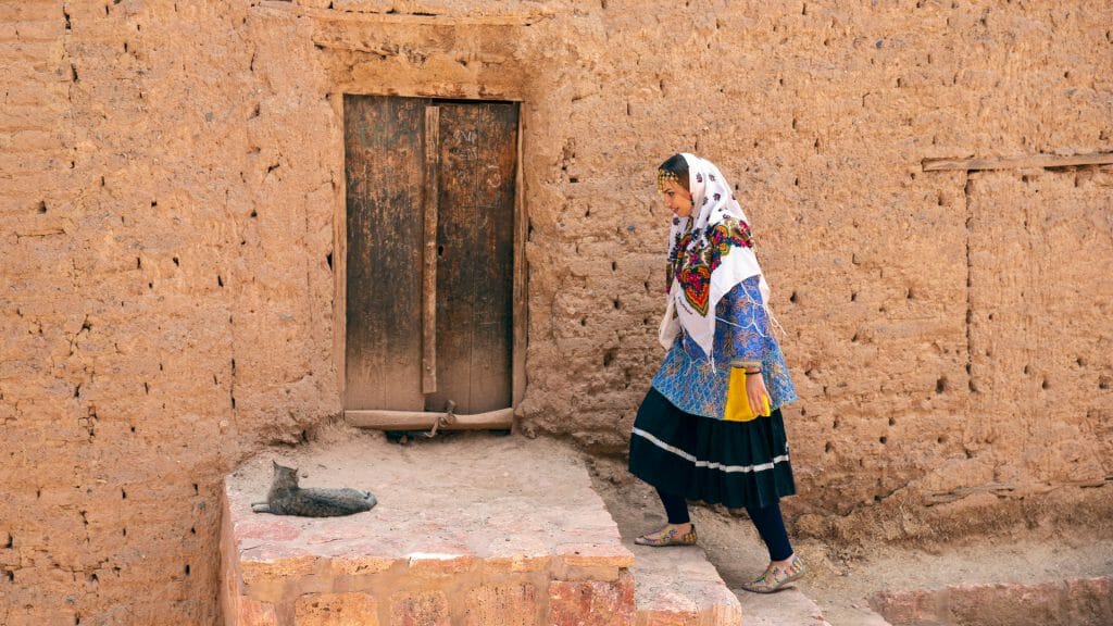 Side of mud house with woman in traditional dress walking up steps to wooden door.