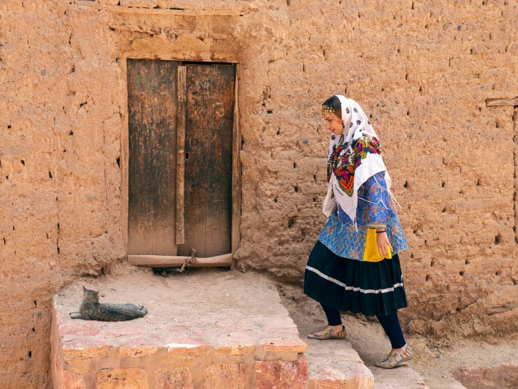 Side of mud house with woman in traditional dress walking up steps to wooden door.