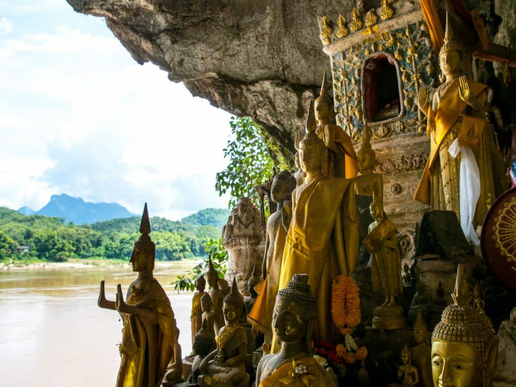 View out from cliffside caves towards river and jungle beyond. Buddhist sculptures within cave in the foreground.