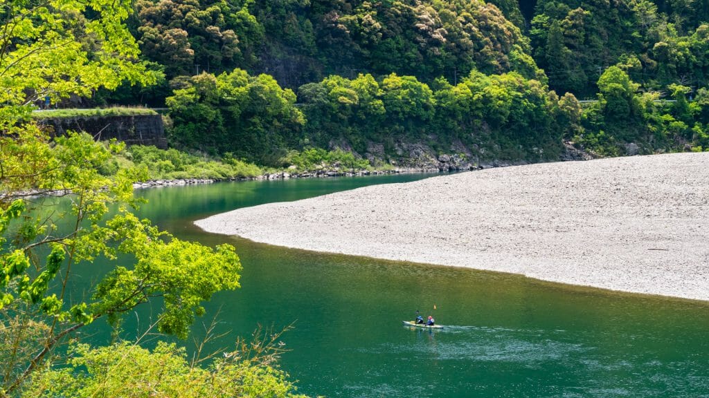 Deep green river with solitary kayaker, shore on right and forest on left.