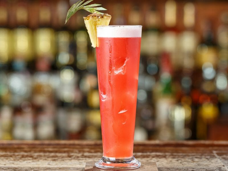 Close up of a singapore sling cocktail, long glass, pink drink with pineapple garnish.