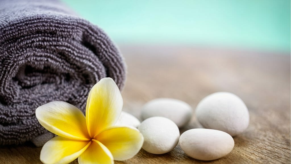 Rolled grey towel with a yellow frangipani flower infront of it and four white stones to one side.