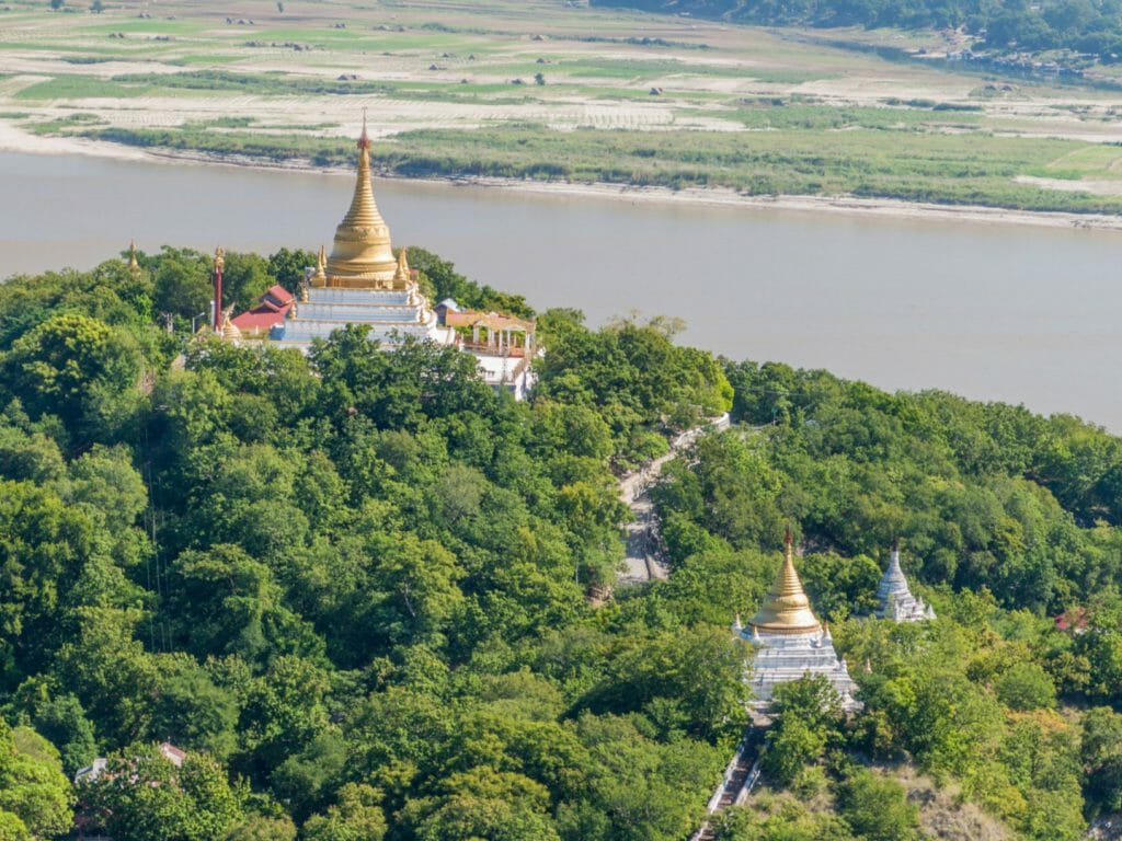 Golden stupas and temples set in lush woodland with river beyond.