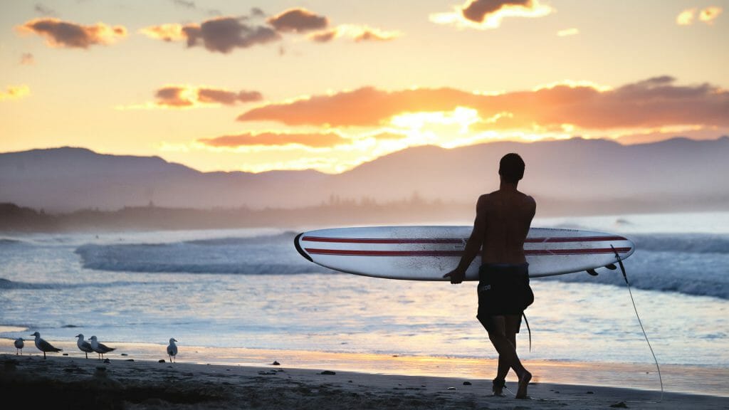 Surfer walking on the shore along the beach looking at the stunning sunset in Byron Bay