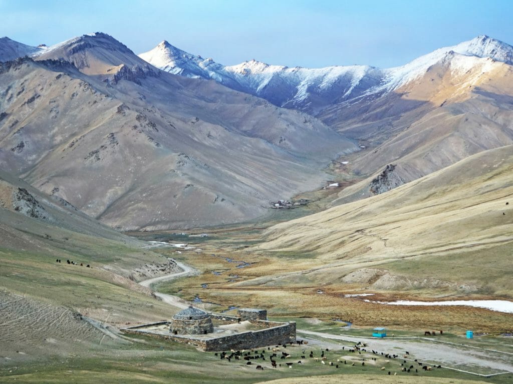 Wide panoramic view valley with caravanserai at the base and snowy peaks in distance.