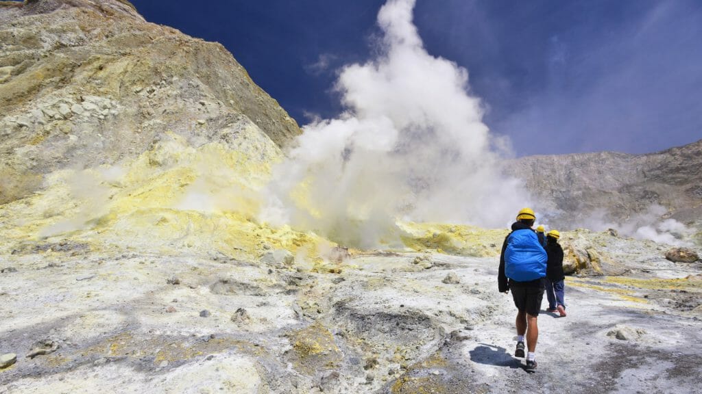 Back of a man with blue rucksac walking on volcanic landscape of white and yellow with smoke rising.