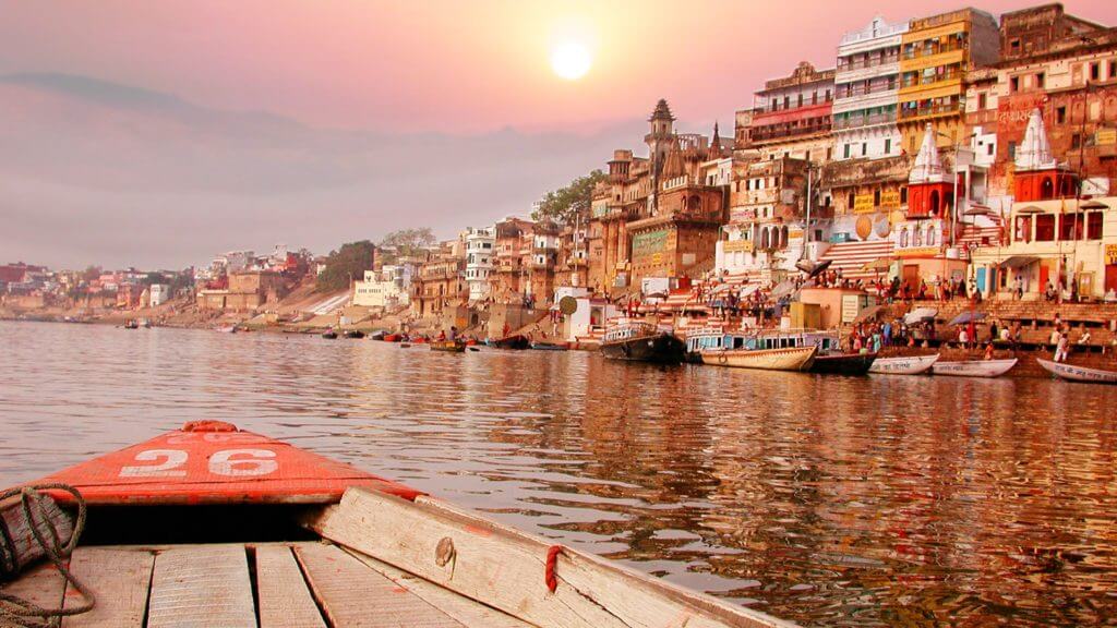 A wooden boat sails on the Ganges River next to a community at sunset