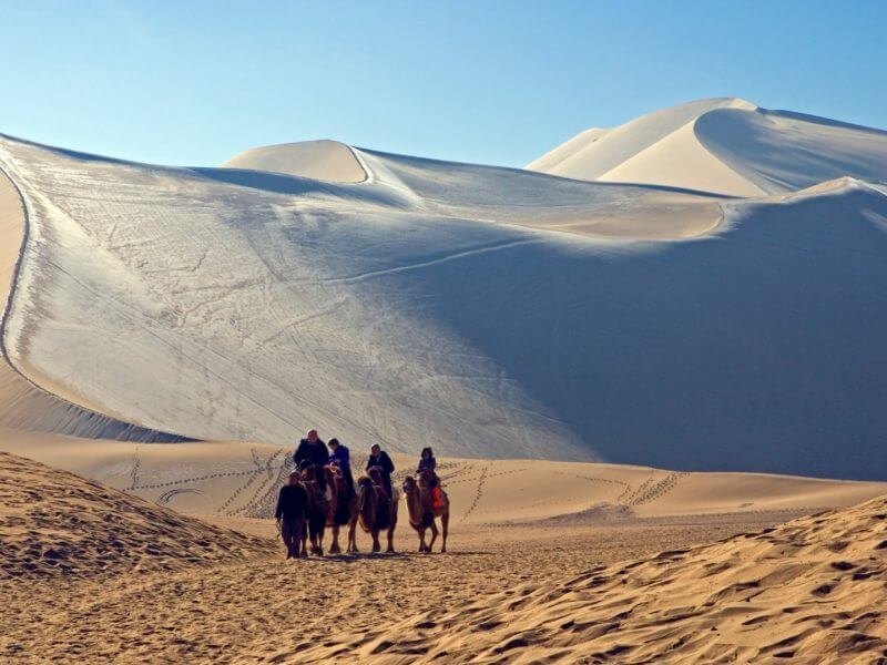 Rolling desert dunes with group of camel riders in foreground.