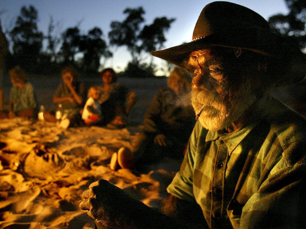 Aboriginal man in hat sitting on sand round campfire at dusk.
