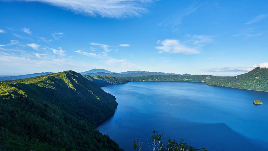 View down onto huge blue lake in a volcanic crater with lush covered crater rim and blue sky.