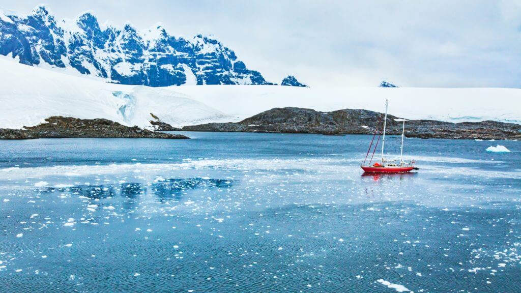 A small red yacht sailing on blue ocean against a backdrop of snow covered mountains in Antarctica