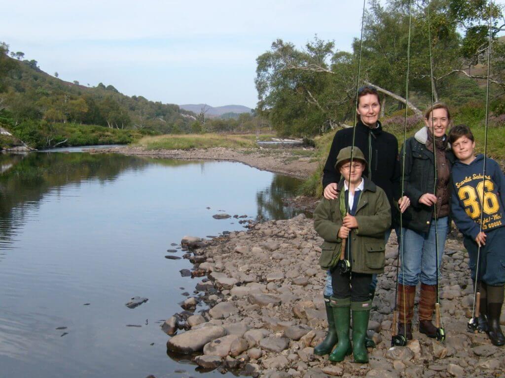 Two women and two boys holding fishing rods standing alongside a river.