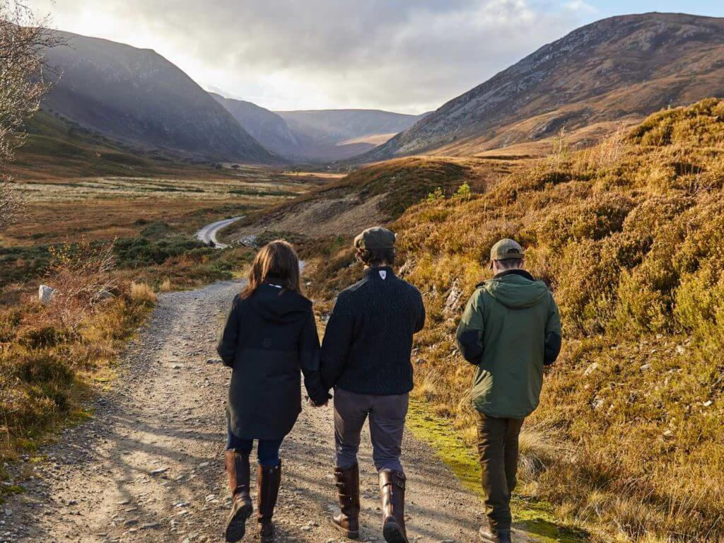 Rear view of three people walking along a track surrounded by autumnal, mountainous landscapes.