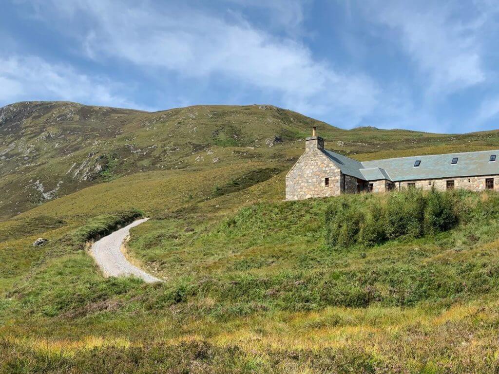 Dirt track leading to stone single storey cottage surrounded by undulating green hills an mountains against blue sky.