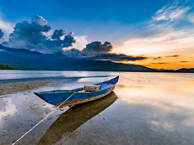 Solitary rowing boat on a lagoon as sun sets over water and mountains in background.