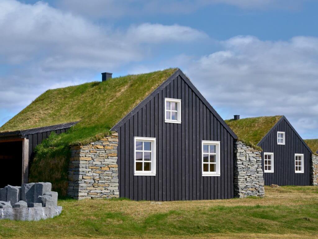 Row of black wooden clad, turf roofed houses of Torfhus Retreat in summer with grass infront and cloudy blue sky.