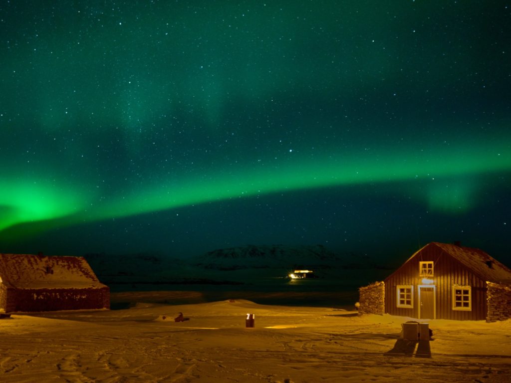 Night shot of wooden clad accommodation at Torfhus Retreat against the northern lights skies the colour of teal and emerald.