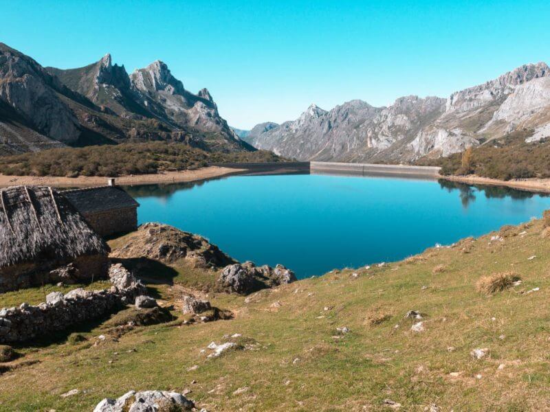 Bright blue glass like lake surrounded by mountains and grassy slopes with thatched shack on shores.