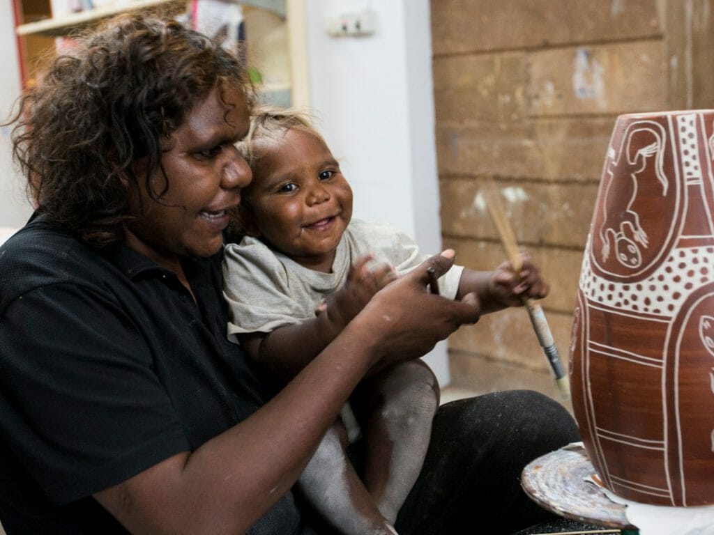 Aboriginal woman with baby son on lap decoorating a pottery vase with traditional dot art.