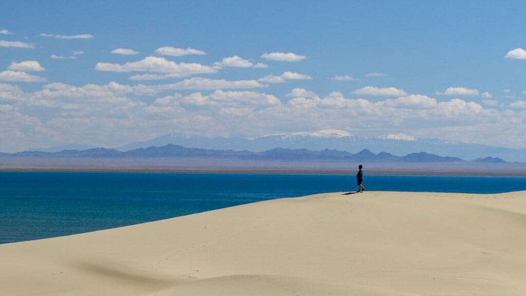 Solitary figure on the crest of a sand dune with a vast blue lake behind and snow capped peaks in the distance.