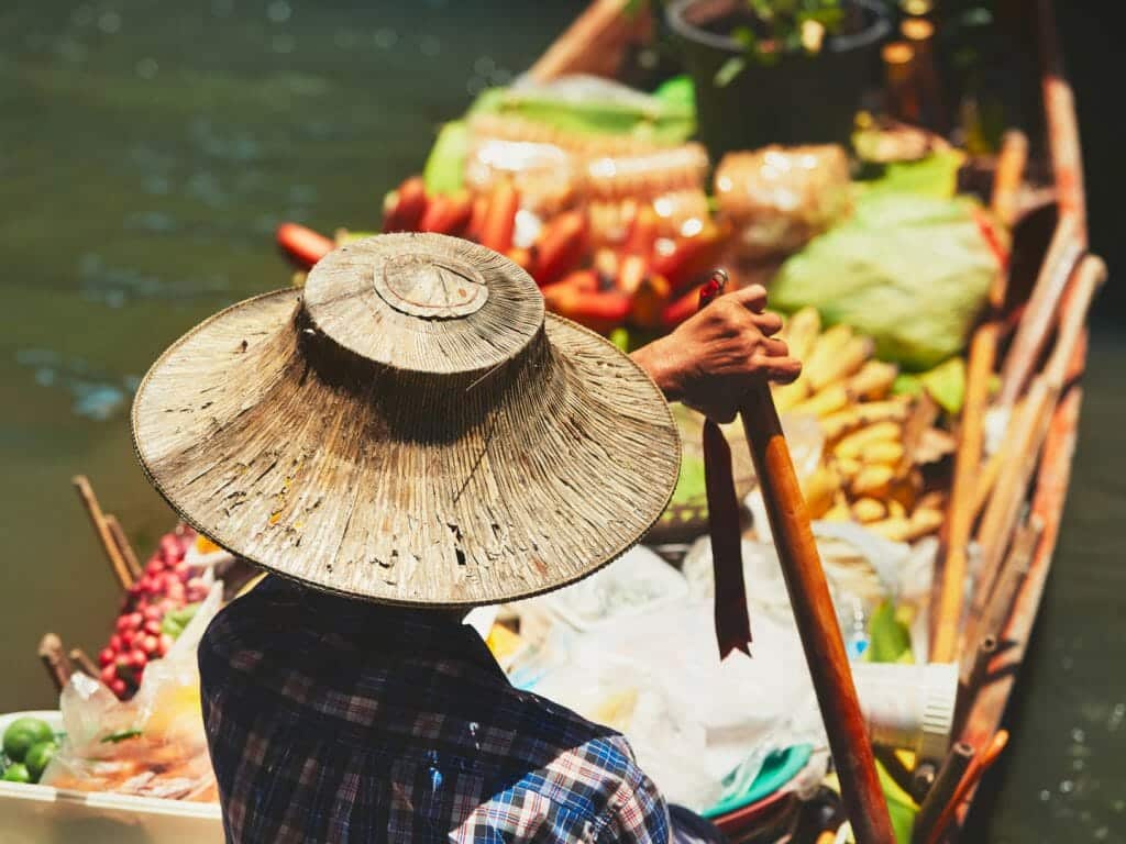 shot from behind of floating market trader wearing traditional wicker hat with boat full of colourful fruit and veg.