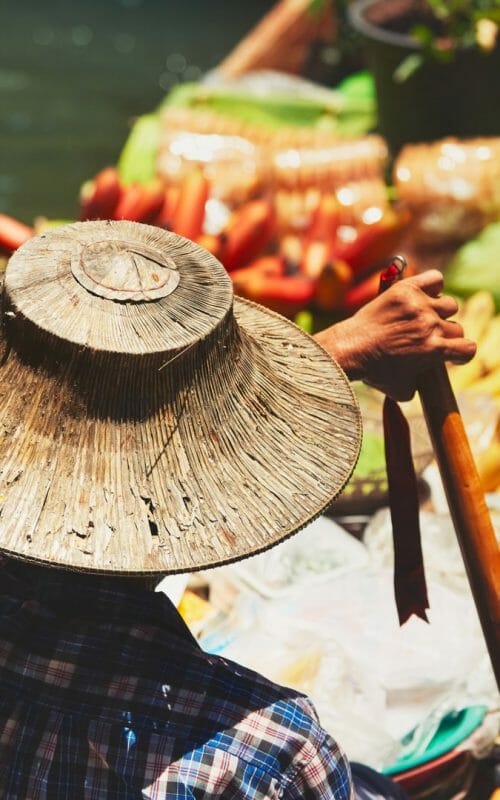 shot from behind of floating market trader wearing traditional wicker hat with boat full of colourful fruit and veg.