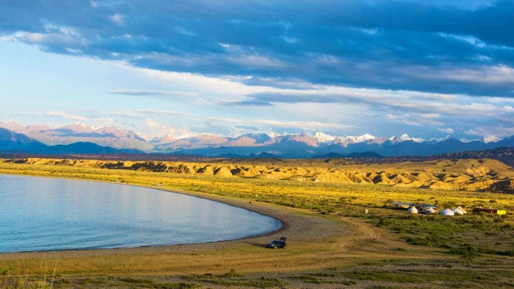 View of lime green grassland leading to shores of curved lake and snowy distant mountains.