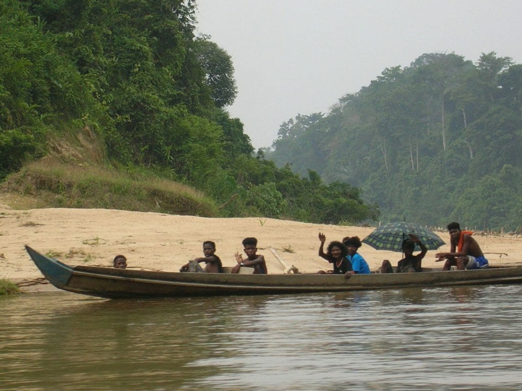 Traditional wooden longboat with group of orang-asli people onboard - on river with sand bank behind and rainforest