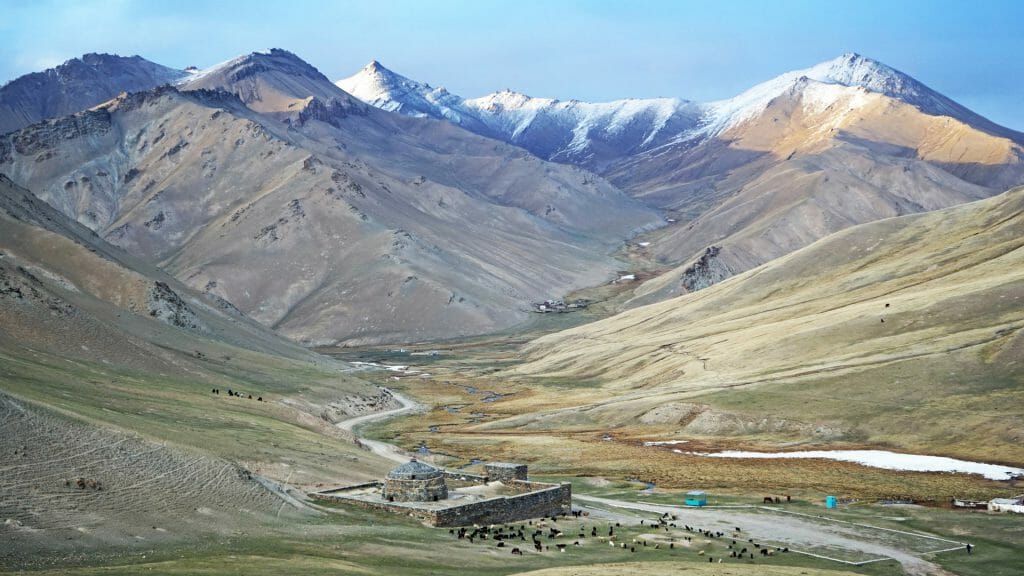 Wide panoramic view valley with caravanserai at the base and snowy peaks in distance.