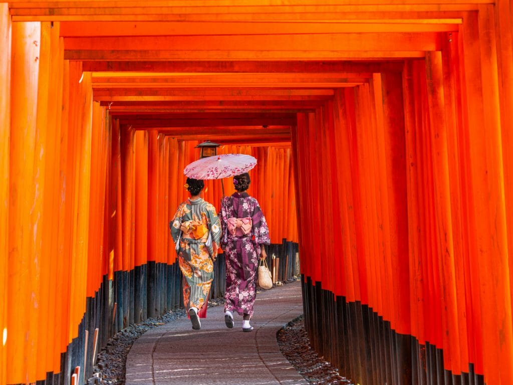 Two women walking away through a square archway of orange pillars.