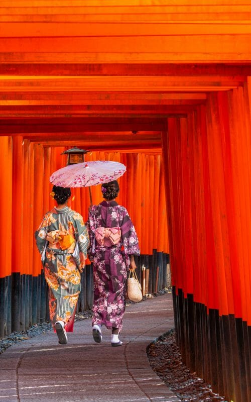 Two women walking away through a square archway of orange pillars.