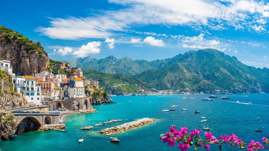 The ancient cliffside town of Atrani overlooks sail boats on turquoise blue ocean on Italy's Amalfi Coast