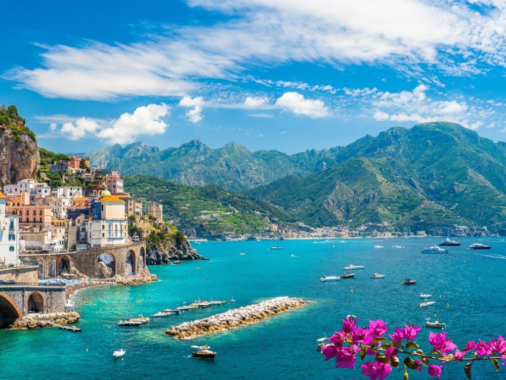 The ancient cliffside town of Atrani overlooks sail boats on turquoise blue ocean on Italy's Amalfi Coast