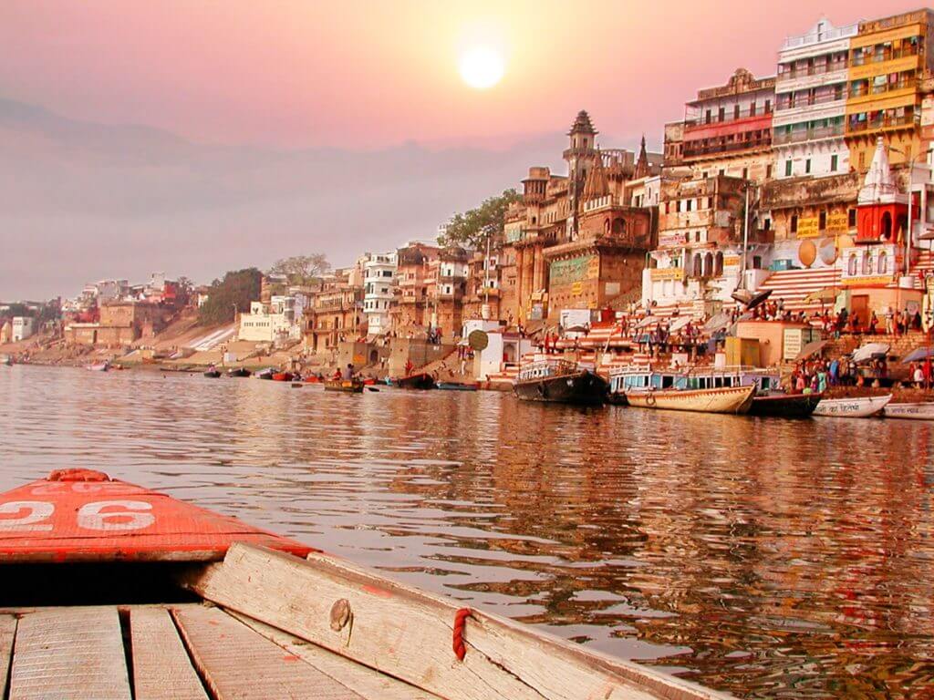A wooden boat sails on the Ganges River next to a community at sunset
