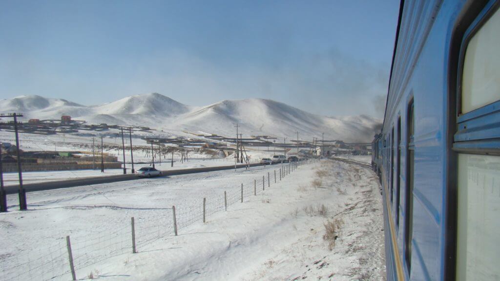 Side of a train travelling through snowy landscapes with snow covered hills in background.