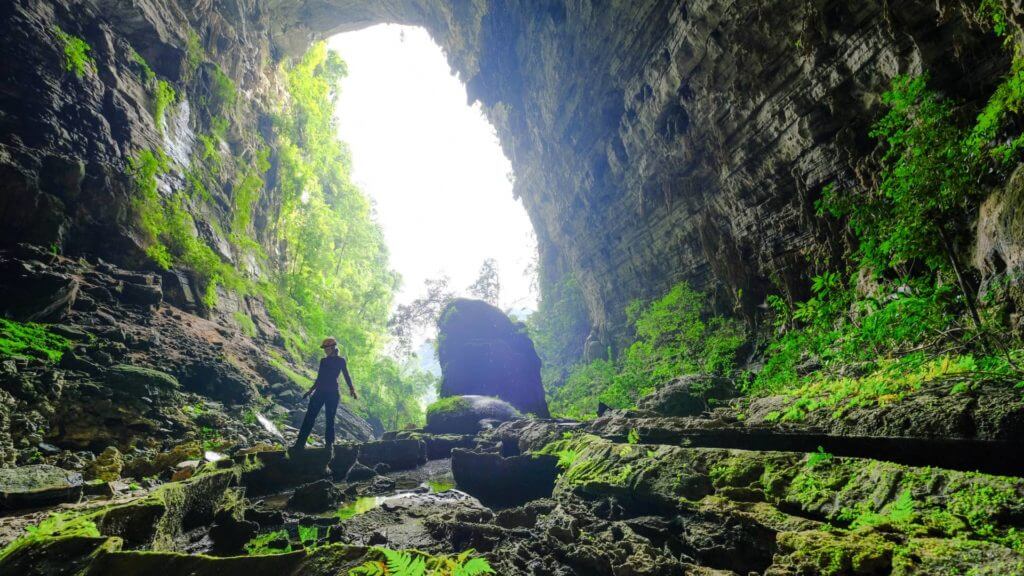 View from interior of rocky cave lined with moss towards cave entrance with silhouette of woman at entrance.